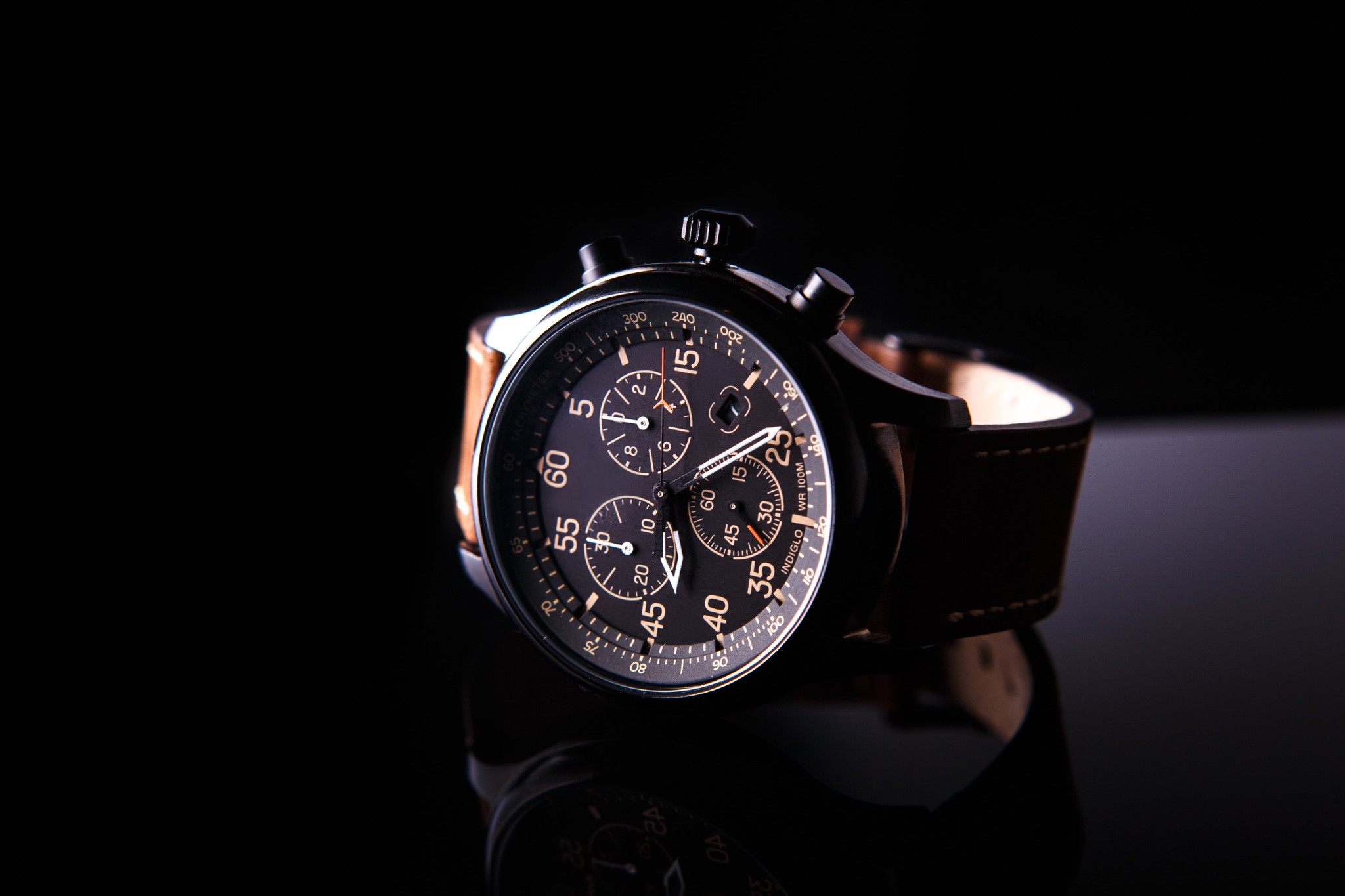 Dark chronograph with a brown leather strap reflected on a black surface